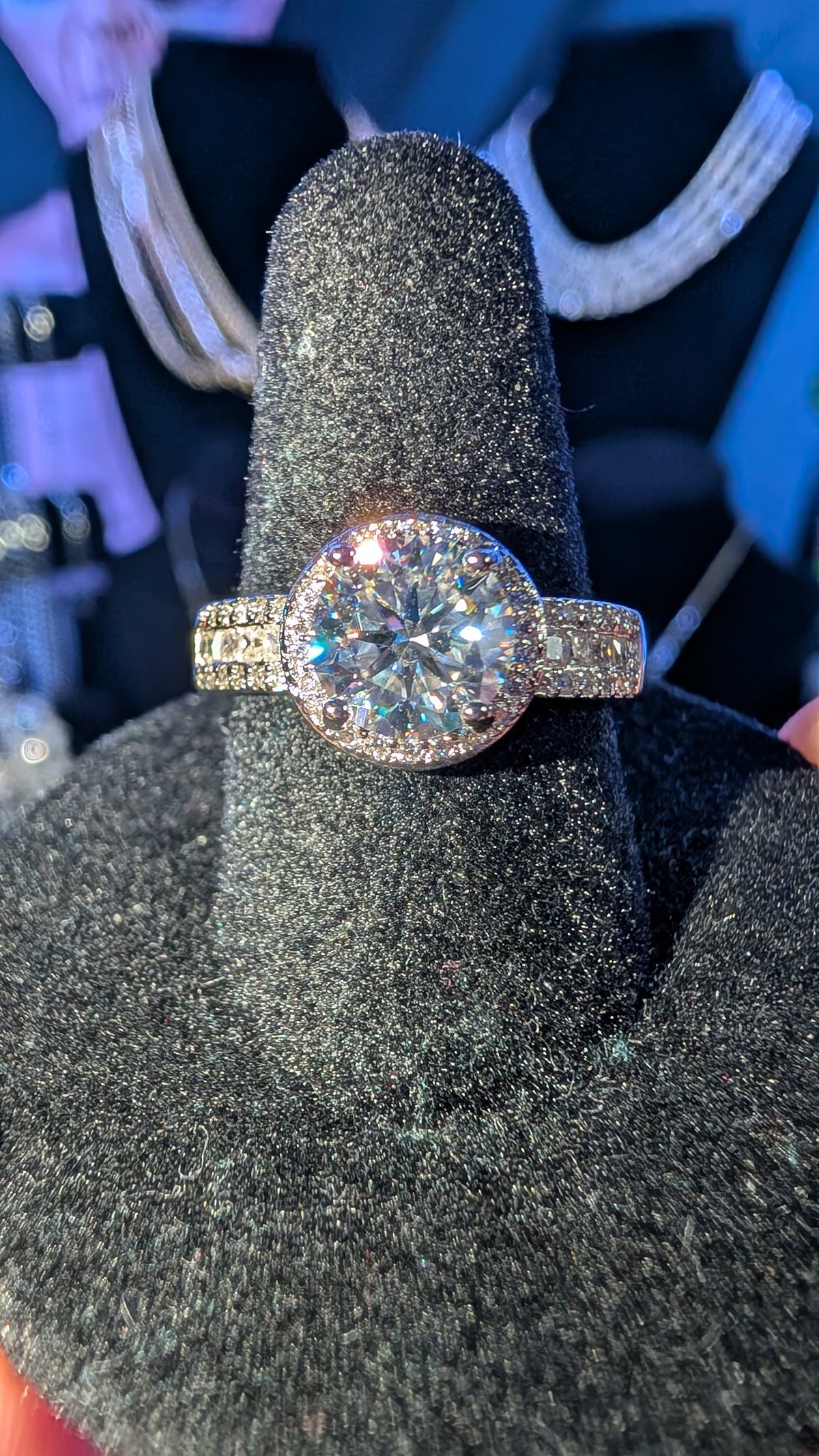 A halo ring with a large round brilliant-cut center stone and pavé-set side stones on a textured black display stand.